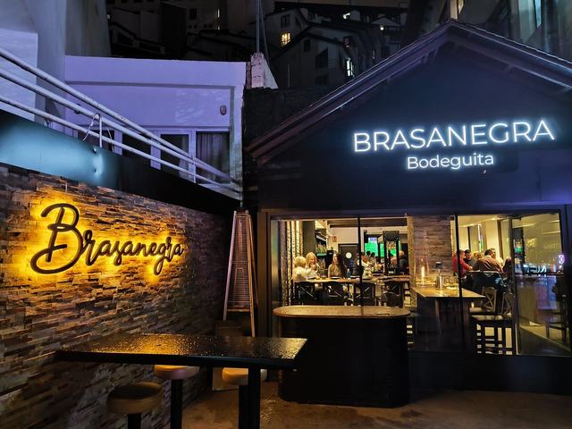 Fachada de Brasanegra Bodeguita de noche, con el letrero iluminado sobre la pared de piedra y el comedor lleno